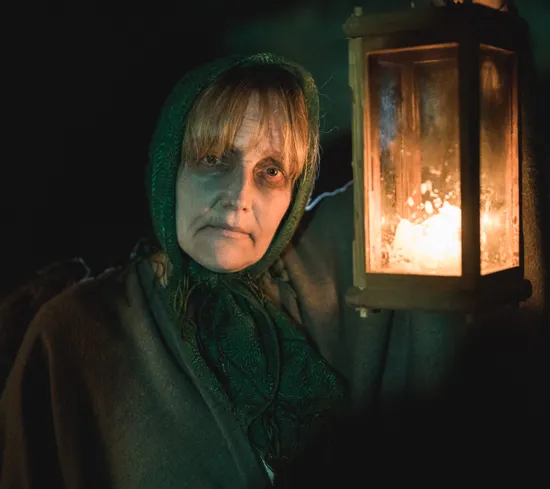 Frau in dunklem Umhang und grünem Kopftuch hält leuchtende Laterne bei schwachem Licht